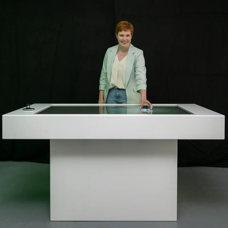 Interaktiver Tisch mit Touchscreen auf dem eine Frau die Benutzung demonstriert. Sie hält einen der interaktiven Bedienungsknöpfe in der Hand und lächelt in die Kamera.