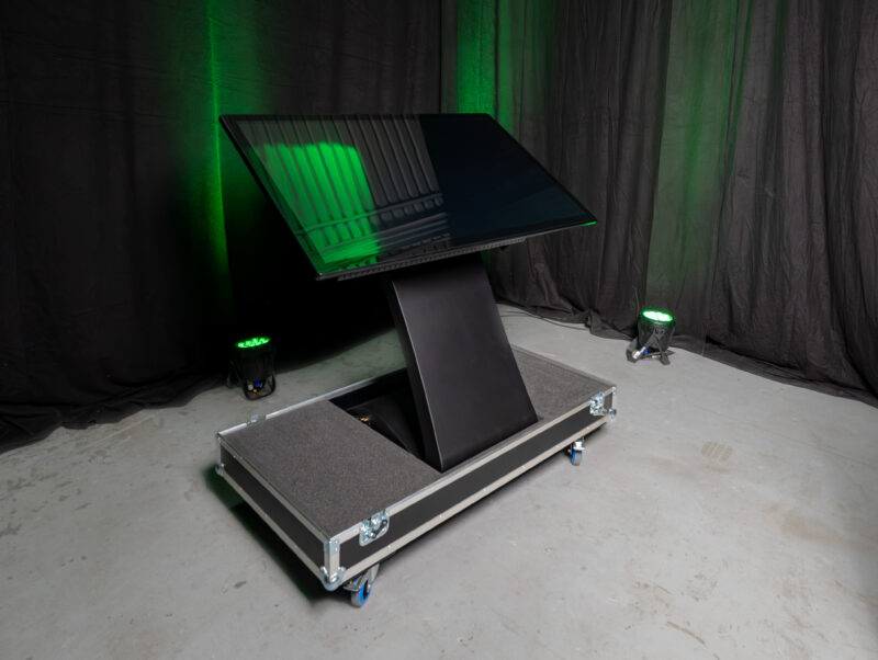 Ein großes, schwarzes interaktives Touch-Terminal mit schräg angewinkeltem Bildschirm steht in der passgenauen Bodenschale eines geöffneten Transport-Cases (Flightcase). Das Terminal befindet sich auf Rollen in einem Raum mit dunklen Vorhängen und grüner Event-Beleuchtung.