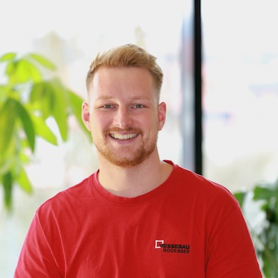 Porträtaufnahme von Fabian Bucher, Projektleiter bei Messebau Bodensee. Er blickt freundlich und lächelnd in die Kamera, hat kurze rot-blonde Haare und einen Bart. Er trägt ein rotes T-Shirt mit dem Logo von Messebau Bodensee auf der linken Brust. Der Hintergrund ist hell und leicht unscharf mit grünen Pflanzen im Anschnitt.