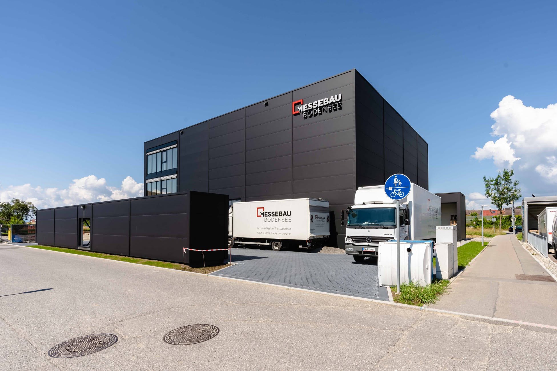 Außenansicht des modernen, anthrazitfarbenen Firmengebäudes der Messebau Bodensee Volk GmbH. An der Fassade ist das Firmenlogo angebracht. Auf dem gepflasterten Hof vor dem Gebäude stehen zwei weiße LKWs für den Logistiktransport, einer davon ebenfalls mit dem Firmenlogo bedruckt. Im Vordergrund verläuft eine Straße bei klarem, leicht bewölktem Himmel.