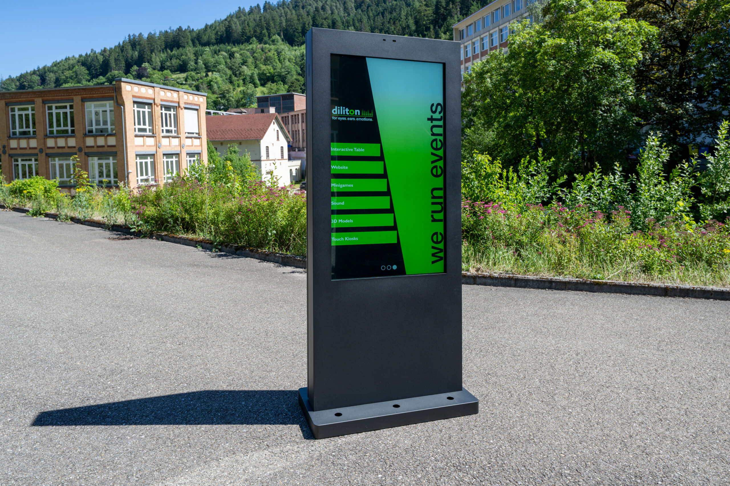 Das Foto zeigt eine freistehende, anthrazitfarbene Informationsstele aus Metall auf einem asphaltierten Platz bei Sonnenschein. Das vertikale Display zeigt ein grün-schwarzes Design mit dem Firmennamen „diliton GmbH“ und dem vertikalen Schriftzug „we run events“. Links stehen grüne Menüschaltflächen, unter anderem beschriftet mit „Interactive Table“, „Website“, „Minigames“, „Sound“ und „3D Models“. Im Hintergrund sind ein gelbes Fabrikgebäude mit vielen Fenstern, üppige grüne Büsche mit rosa Blüten, bewaldete Hügel und ein wolkenloser, blauer Himmel zu sehen. Die Stele wirft einen langen, ausgeprägten Schatten nach links vorne auf den Asphalt.