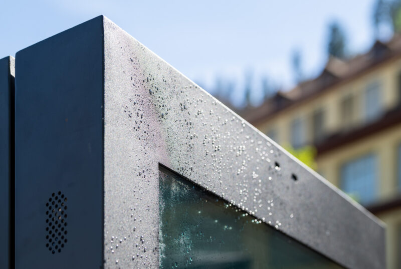 Detailaufnahme der oberen Ecke eines digitalen Outdoor-Kundenstoppers im Sonnenlicht mit Wassertropfen auf dem Rahmen. An der linken Gehäuseseite sind die integrierten, wetterfesten Lautsprecher-Öffnungen sichtbar.