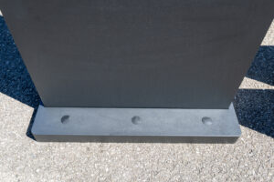 Nahaufnahme vom massiven, anthrazitfarbenen Standfuß einer digitalen Outdoor-Touchstehle auf Asphalt mit drei Abdeckungen an der Vorderseite.