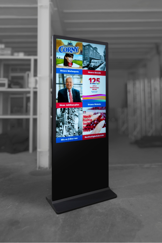 Eine freistehende, schwarze digitale Infostele in einem Raum, dessen Hintergrund in Schwarz-Weiß gehalten ist. Der farbige, vertikale Bildschirm zeigt eine Kachel-Übersicht der Schwartauer Werke mit Inhalten wie „Unsere Werbespots“ (mit Corny-Logo), „Unsere Chronik“, „Unser Jubiläumsfilm“, „Unsere Website“ (mit dem 125-Jahre-Logo), „Wie es früher war“ und dem „Nachhaltigkeitsbericht“.