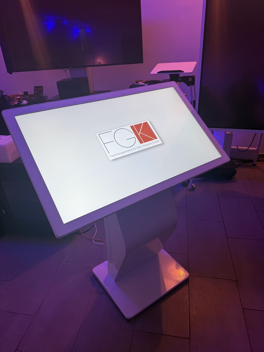 Ein schräg angewinkeltes, interaktives Touch-Terminal in Weiß, auf dessen Bildschirm das Logo des Forschungsinstituts für Glas und Keramik (FGK) zu sehen ist.