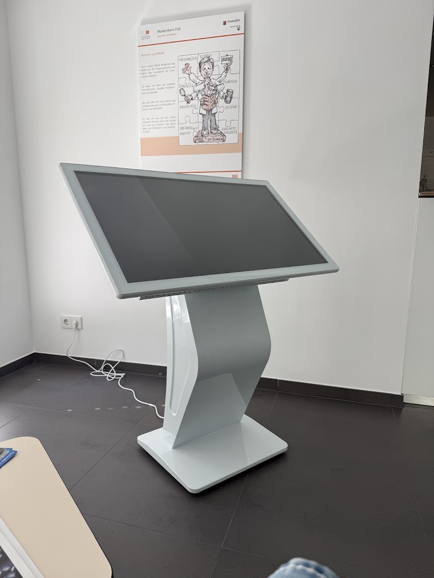 Ein freistehendes, weißes Touch-Terminal mit ausgeschaltetem Bildschirm in einem hellen Raum vor einem Infoplakat zum FGK-Markenkern.