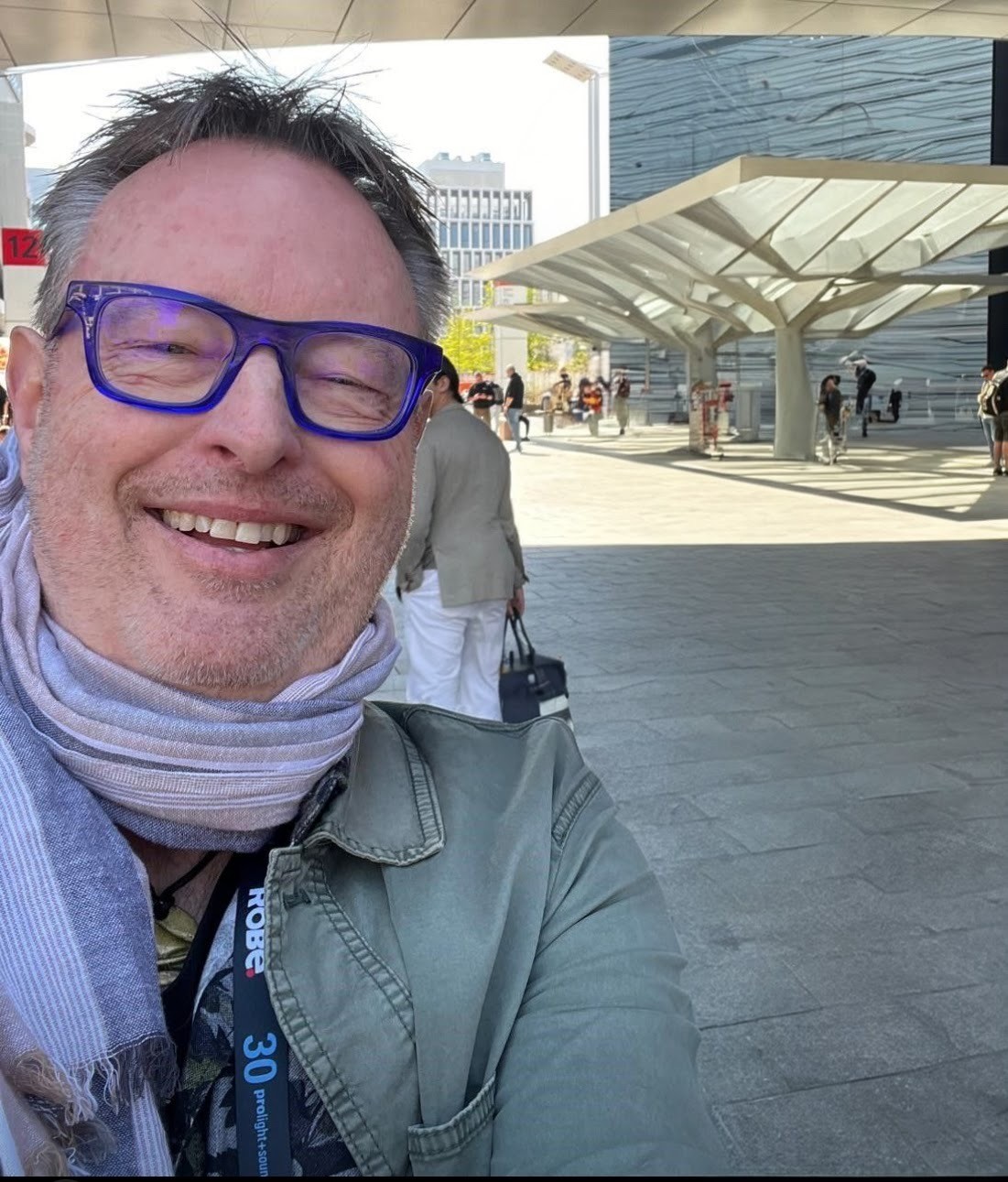 Ein lächelndes Selfie von Mario Klein mit blauer Brille, Schal und einem Messe-Lanyard der „prolight+sound“, im Hintergrund ein moderner Außenbereich mit Messebesuchern.