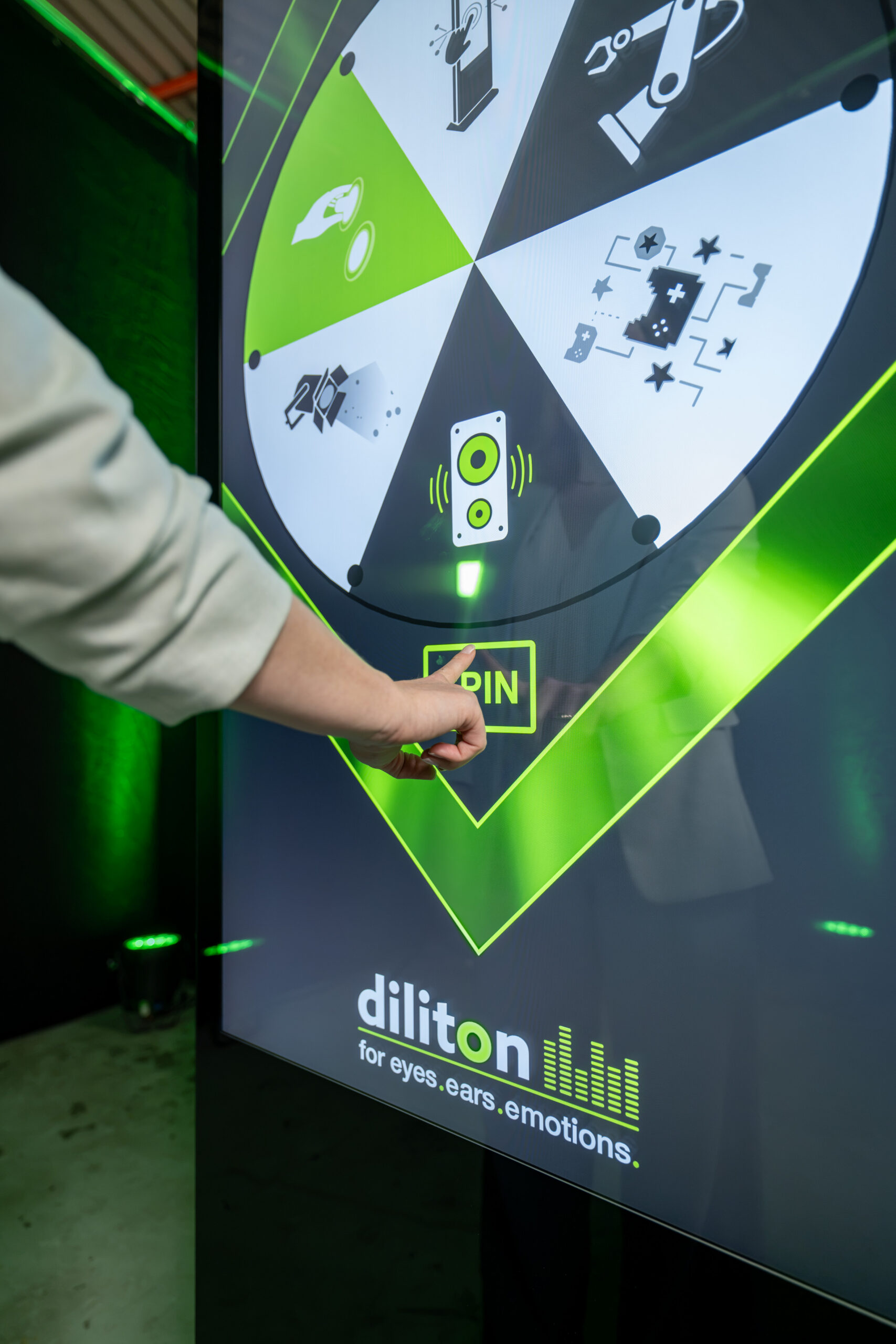 Eine Nahaufnahme zeigt die Hand einer Person, die den gelben „SPIN“-Button auf dem Display einer vertikalen Touch-Stele von diliton drückt. Auf dem Bildschirm läuft eine interaktive Glücksrad-Anwendung in Grün und Weiß, die verschiedene Symbole wie Lautsprecher oder Überwachungstechnik zeigt. Im Hintergrund schimmert ein grüner Lichteffekt im abgedunkelten Raum.