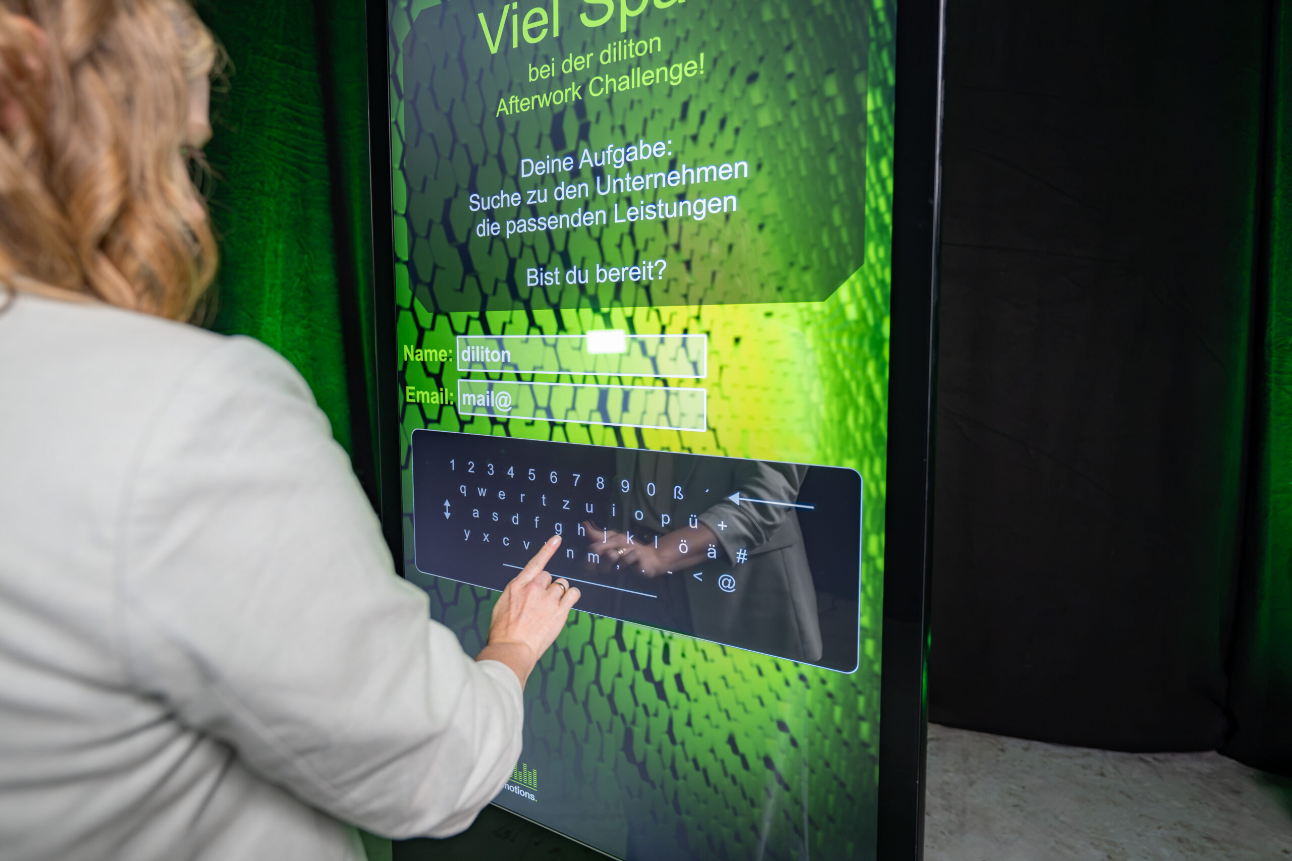 Die Nahaufnahme zeigt eine Frau von hinten links, die nach einem interaktiven Spiel ihre Kontaktdaten zur Leaderfassung auf einer vertikalen Touch-Stele eingibt. Das Display führt durch die „diliton Afterwork Challenge“ und fordert zur Eingabe von Name und E-Mail-Adresse auf. Die Nutzerin tippt gerade über die eingeblendete digitale Bildschirmtastatur, um das Formular auszufüllen.