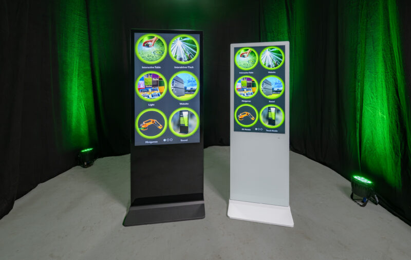 Das Bild zeigt zwei digitale Touchstelen im direkten Vergleich: ein Modell im schwarzen und ein Modell im weißen Metallgehäuse. Beide Displays zeigen interaktive Software-Inhalte und verdeutlichen die flexiblen farblichen Gestaltungsmöglichkeiten für den optimalen Markenauftritt.
