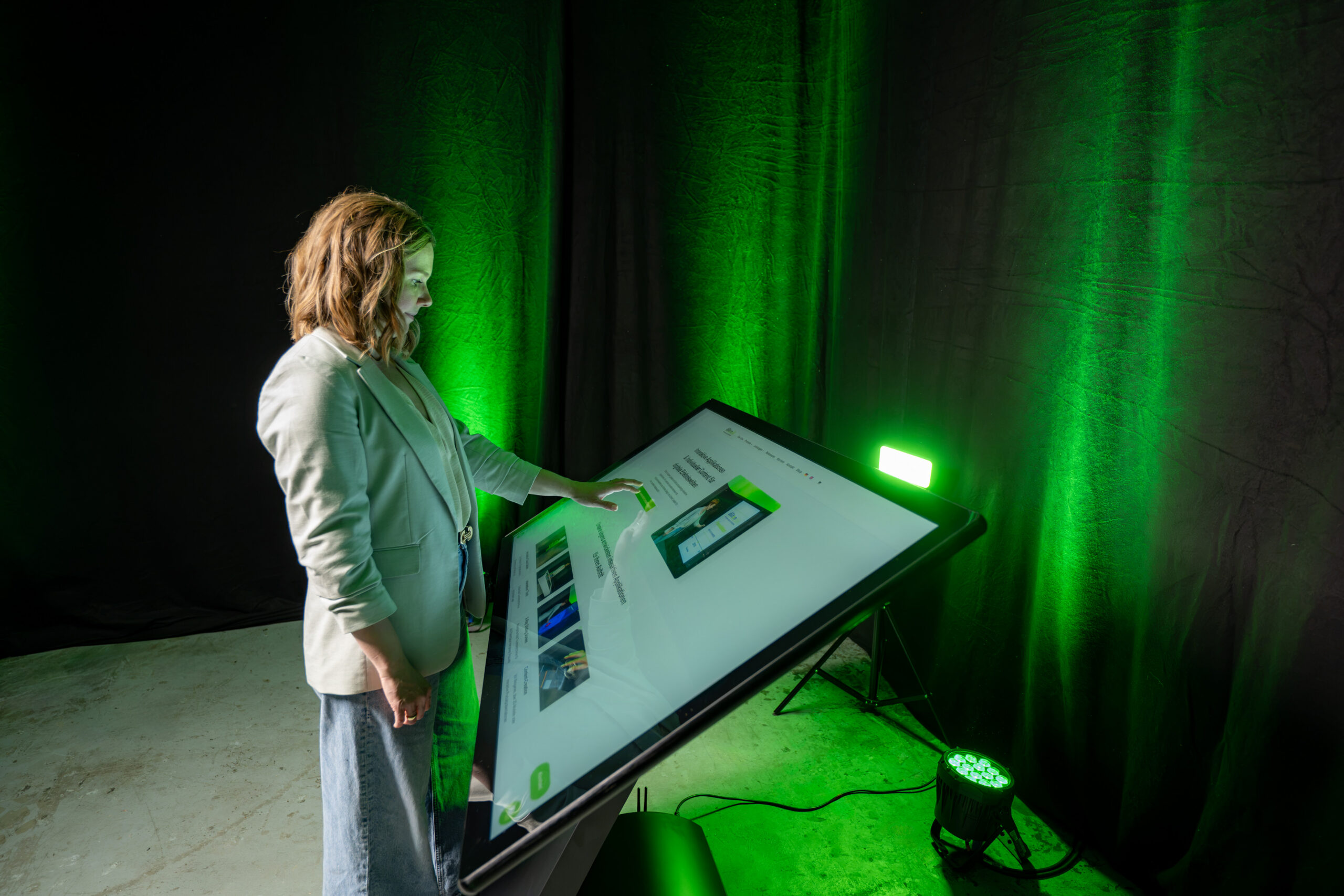 Perspektivische Seitenansicht einer Frau im hellen Blazer, die per Touch-Geste eine Webseite auf dem großen, horizontal ausgerichteten Bildschirm einer Kiosk-Stele bedient.