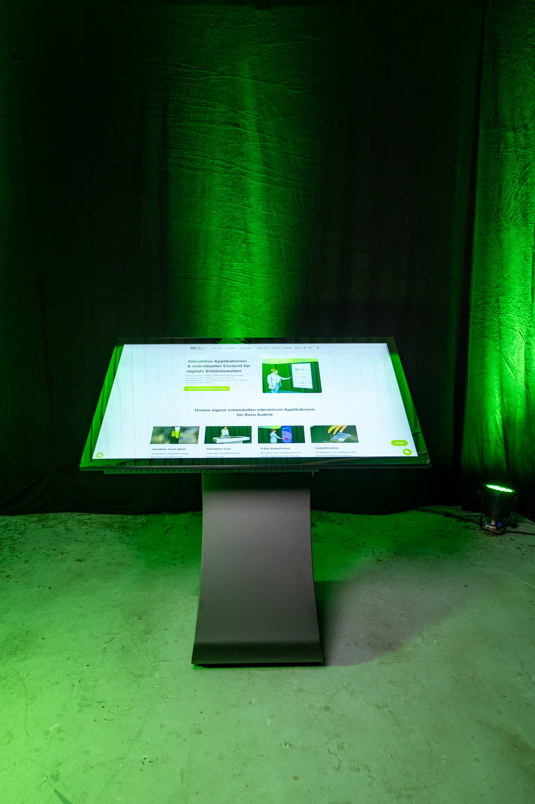 Frontale Studioaufnahme im Hochformat von einem 55 Zoll großen, horizontal ausgerichteten Touch Terminal mit geschwungenem S-Sockel Standfuß und der diliton Website auf dem Display.