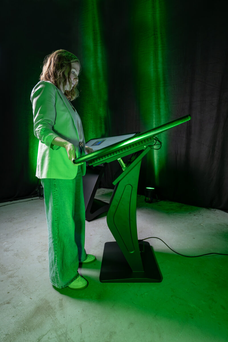 Detailaufnahme einer Hand, die das Gehäuse einer horizontalen Kiosk-Stele anhebt, um die flexible und ergonomische Neigungsverstellung des Touch-Displays individuell anzupassen.