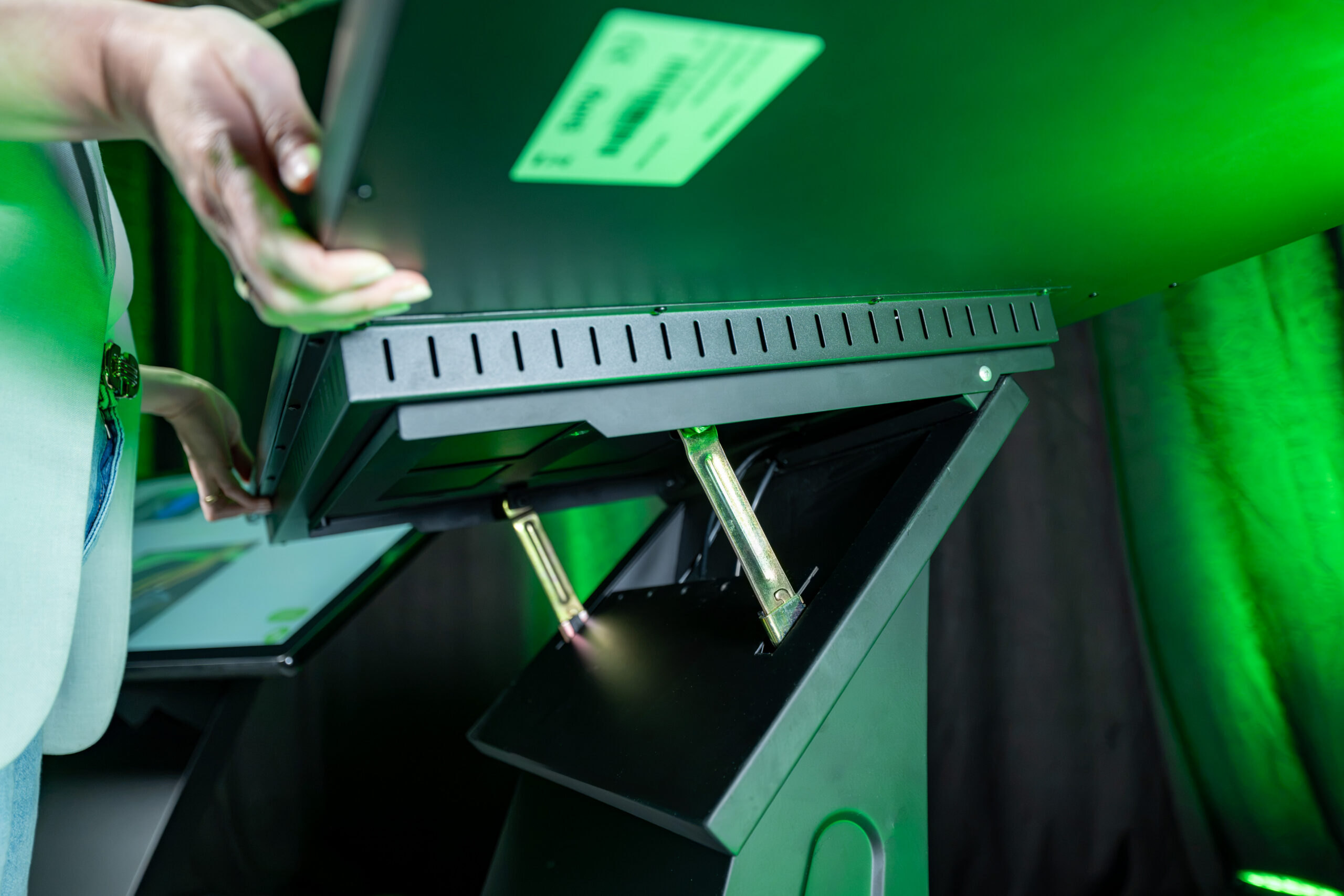 Technische Detailaufnahme der Unterseite einer Kiosk-Stele: Eine Person verstellt den Neigungswinkel des horizontalen Bildschirms über eine metallische, mehrstufige Raster-Mechanik über dem schwarzen Standfuß.