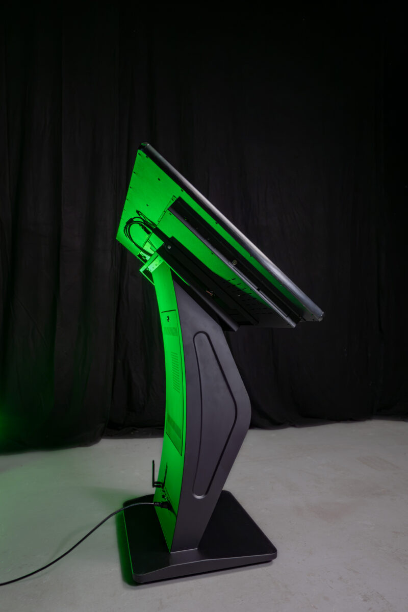 Seitenansicht eines interaktiven Touch Terminals im Querformat mit einem massiven, funktionalen K-Sockel Standfuß in Schwarz vor dunklem Hintergrund.