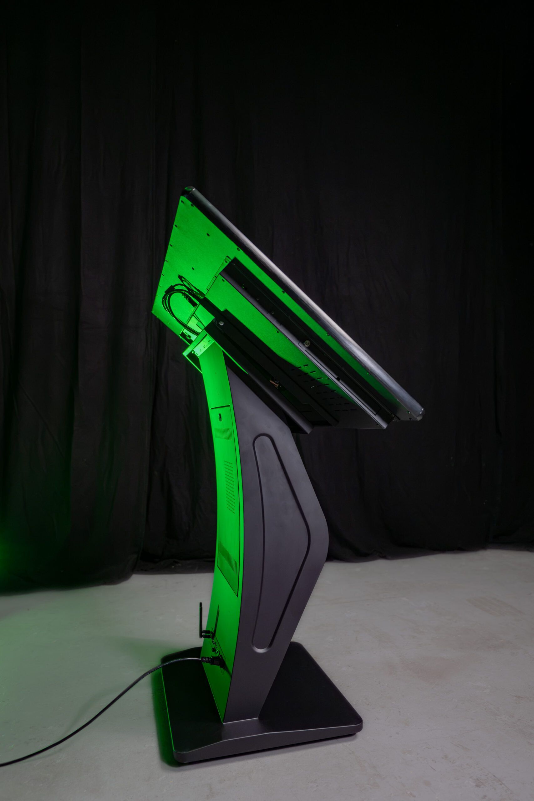 Seitenansicht eines interaktiven Touch Terminals im Querformat mit einem massiven, funktionalen K-Sockel Standfuß in Schwarz vor dunklem Hintergrund.