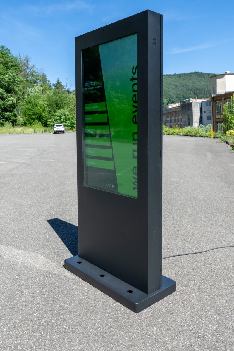 Das Bild zeigt eine vertikale Outdoor-Touch-Stele in einer leicht schrägen Seitenansicht. Das robuste, mattschwarze Metallgehäuse steht standsicher auf einem massiven Sockel im Freien auf einem asphaltierten Platz. Auf dem spiegelungsarmen Display ist eine grüne Benutzeroberfläche der Marke diliton mit der Aufschrift „we run events“ zu sehen. Ein Stromkabel wird flach nach hinten über den Boden weggeführt. Im Hintergrund erstrecken sich grüne Bäume, ein bewaldeter Hügel sowie Fabrikgebäude unter einem klaren, blauen Himmel bei strahlendem Sonnenschein.