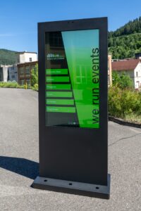 Das Foto zeigt eine freistehende, anthrazitfarbene digitale outdoor Stele aus Metall auf einem asphaltierten Platz bei Sonnenschein. Das vertikale, helle Display zeigt ein grün-schwarzes Design mit dem Firmennamen „diliton“, dem vertikalen Slogan „we run events“ und einer Liste von Menüpunkten. Im Hintergrund sind grüne, bewaldete Hügel, vereinzelte Autos und ältere Industriegebäude unter blauem Himmel zu sehen. Das Gerät wirft einen deutlichen Schatten nach links vorne.