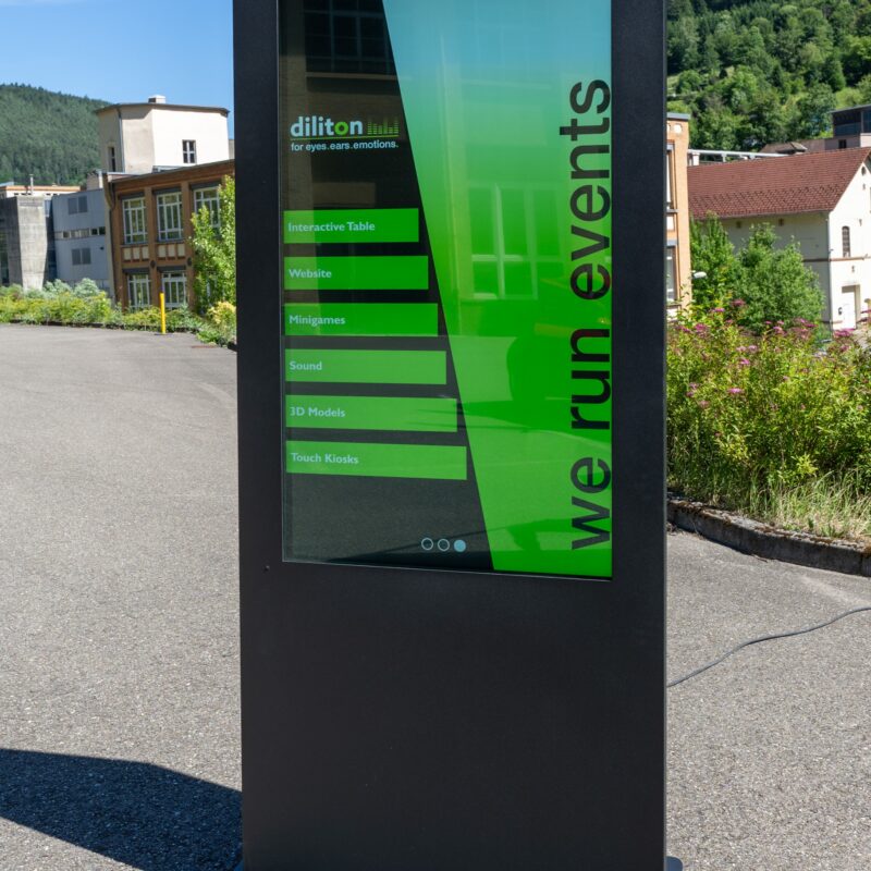 Das Foto zeigt eine freistehende, anthrazitfarbene digitale outdoor Stele aus Metall auf einem asphaltierten Platz bei Sonnenschein. Das vertikale, helle Display zeigt ein grün-schwarzes Design mit dem Firmennamen „diliton“, dem vertikalen Slogan „we run events“ und einer Liste von Menüpunkten. Im Hintergrund sind grüne, bewaldete Hügel, vereinzelte Autos und ältere Industriegebäude unter blauem Himmel zu sehen. Das Gerät wirft einen deutlichen Schatten nach links vorne.