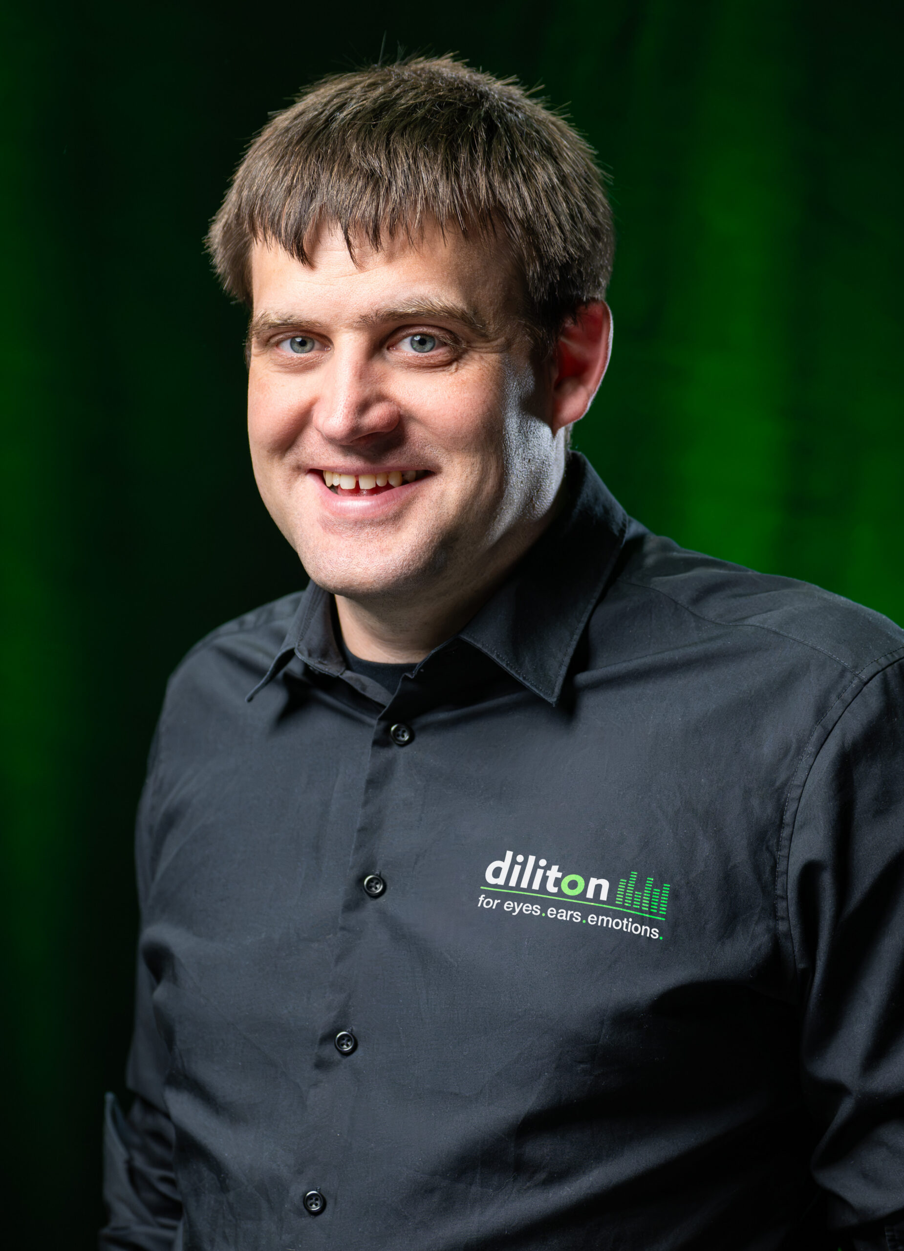 Porträt des Geschäftsführers der diliton GmbH vor einem dunkelgrünen, strukturierten Hintergrund. Er hat kurzes, braunes Haar, blickt lächelnd direkt in die Kamera und trägt ein schwarzes Hemd mit dem Firmenlogo „diliton“ und dem Slogan „for eyes.ears.emotions.“ auf der linken Brustseite.
