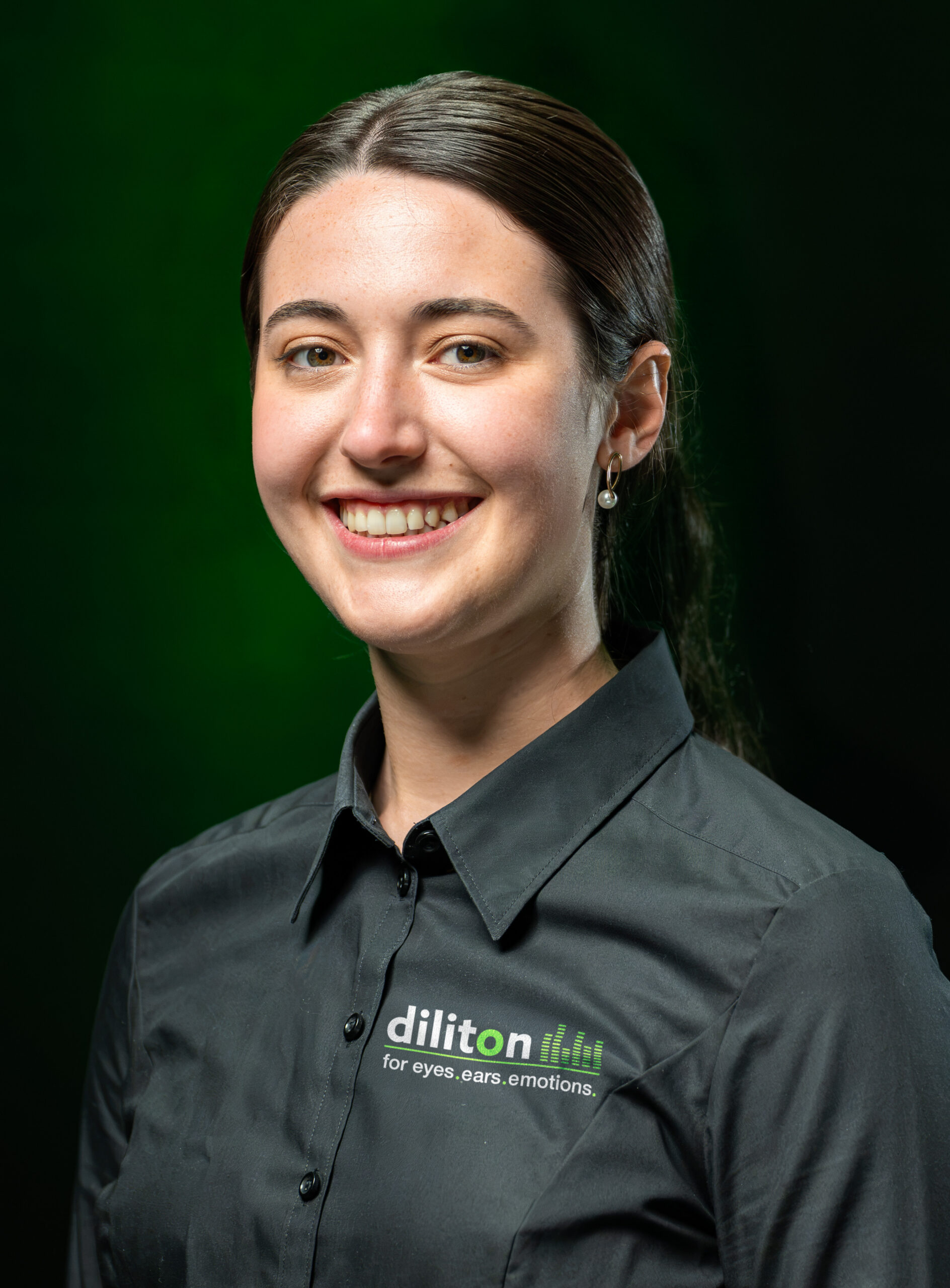 Porträt von Lara Burger, Growth Marketing & Sales Managerin bei der diliton GmbH, vor einem dunkelgrünen Hintergrund. Die junge Frau mit nach hinten gebundenen, dunklen Haaren blickt lächelnd direkt in die Kamera. Sie trägt dezente Ohrringe mit Perlen sowie ein schwarzes Firmenhemd, auf dessen linker Brust das diliton-Logo mit dem Slogan „for eyes.ears.emotions.“ zu sehen ist.