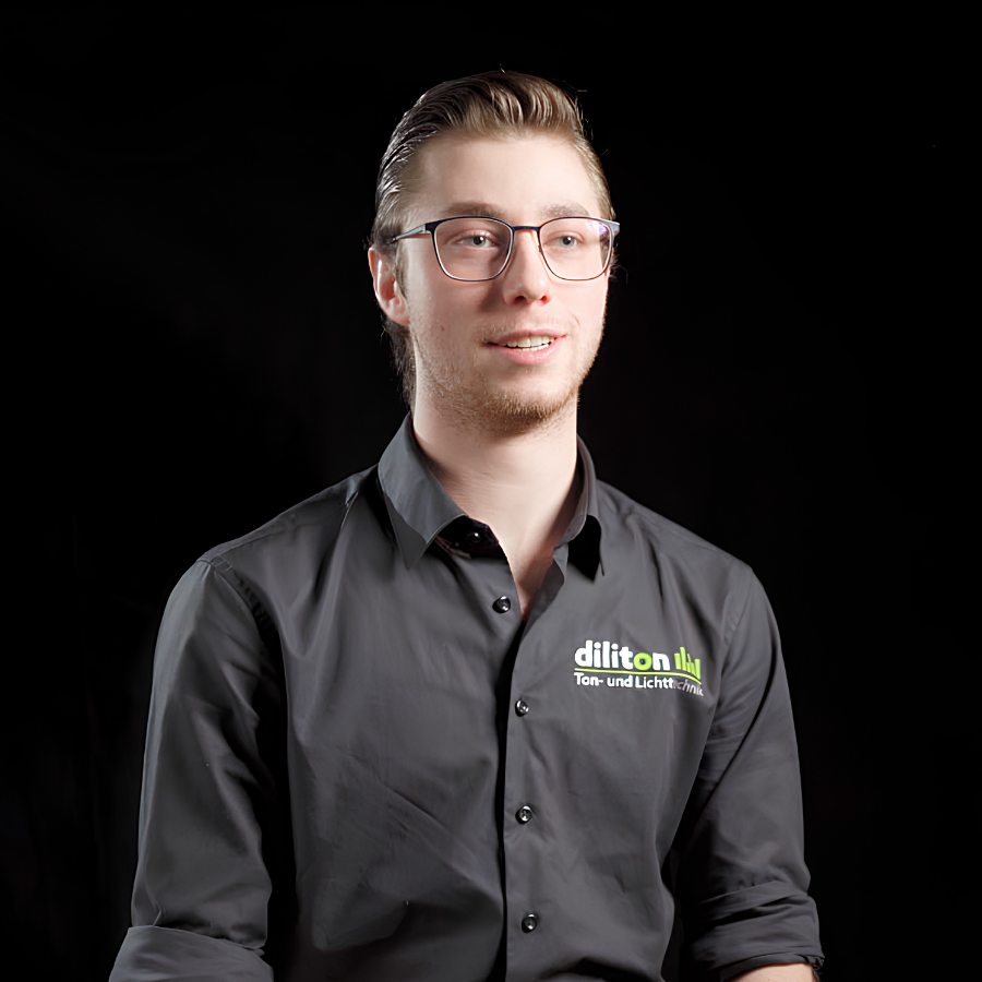 Ein Porträt von Adrian, freiberuflicher Experte für Event-Tontechnik und Sounddesign bei der diliton GmbH, vor einem schwarzen Hintergrund. Der junge Mann mit Brille und zurückgegelten, dunklen Haaren blickt leicht nach rechts an der Kamera vorbei. Er trägt ein dunkelgraues Firmenhemd, auf dem das Logo „diliton“ mit dem Zusatz „Ton- und Lichttechnik“ auf der linken Brust aufgedruckt ist.