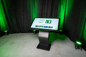 Ein horizontales, leicht geneigtes Touch-Terminal mit einem stabilen, schwarzen K-Sockel steht auf einem Betonboden. Der Hintergrund besteht aus einem schwarzen Vorhang, der von grünen Scheinwerfern angestrahlt wird. Auf dem großen Display ist eine helle Website zu sehen.