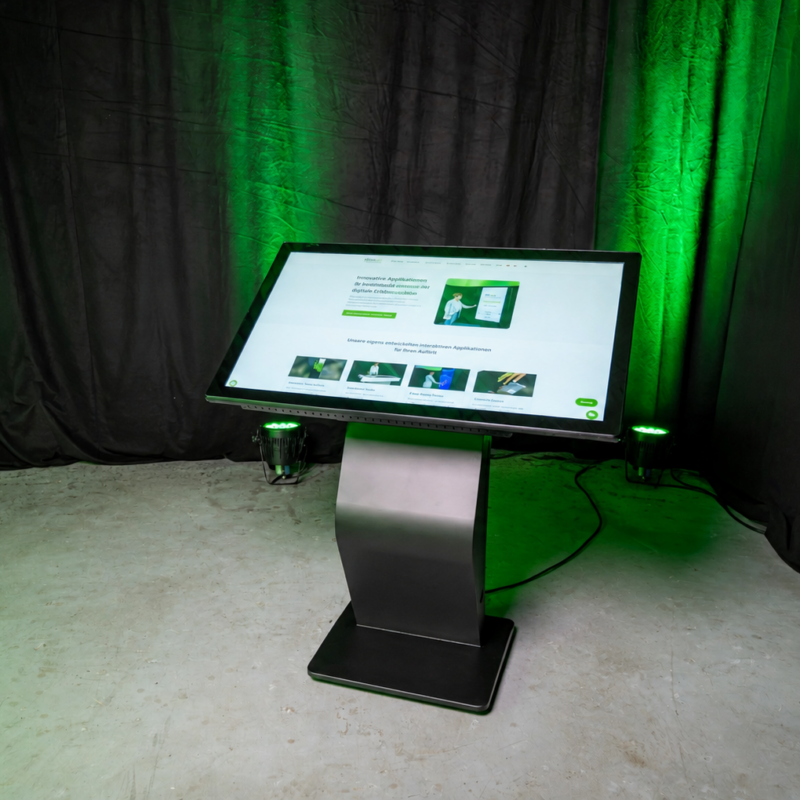 Ein horizontales, leicht geneigtes Touch-Terminal mit einem stabilen, schwarzen K-Sockel steht auf einem Betonboden. Der Hintergrund besteht aus einem schwarzen Vorhang, der von grünen Scheinwerfern angestrahlt wird. Auf dem großen Display ist eine helle Website zu sehen.