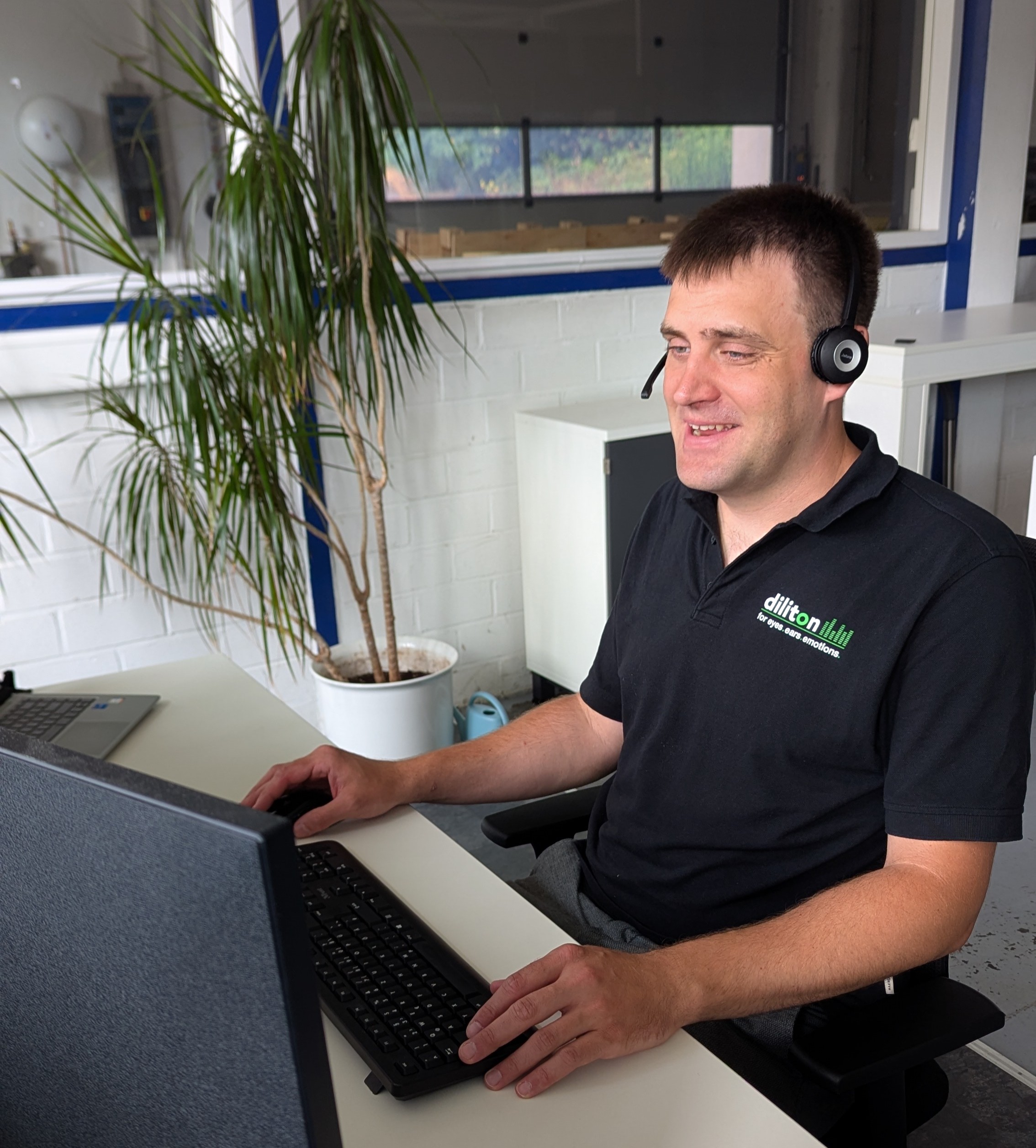 Dennis Dieterle, Geschäftsführer der diliton GmbH, sitzt lächelnd an einem weißen Schreibtisch und blickt auf einen großen PC-Bildschirm im Vordergrund. Er trägt ein schwarzes Poloshirt mit dem grün-weißen Firmenlogo und ein Headset mit Mikrofon am Ohr. Seine Hände liegen auf der Tastatur und der Computermaus. Im hellen Bürohintergrund stehen eine weiße Wand, eine große Zimmerpflanze im Topf und ein Aktenlaufschrank vor einer Fensterfront.