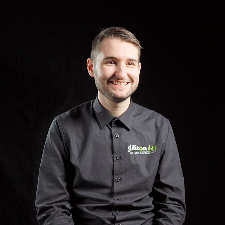 Porträt von Elias, Veranstaltungstechniker mit Fokus auf Lichttechnik bei der diliton GmbH, vor einem schwarzen Hintergrund. Der junge Mann mit kurzen, dunkelblonden Haaren und einem Dreitagebart blickt lächelnd leicht zur Seite. Er trägt ein dunkelgraues Firmenhemd, auf dem das Logo „diliton“ mit dem Zusatz „Ton- und Lichttechnik“ auf der linken Brust aufgedruckt ist.