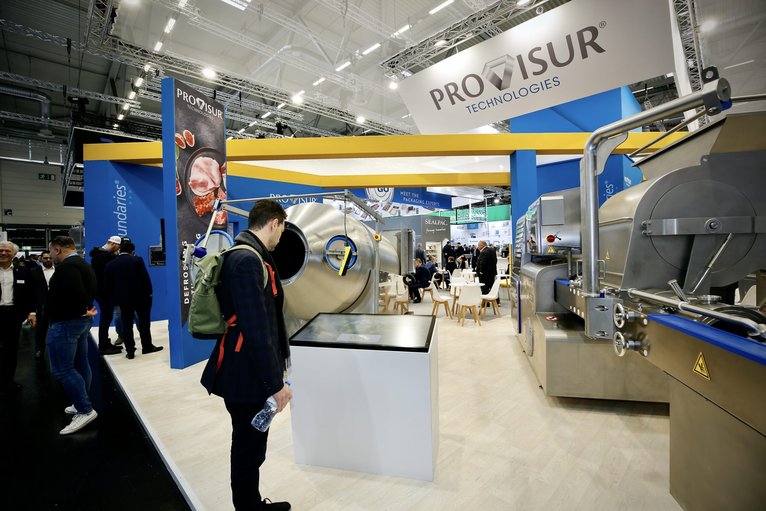 Ein Besucher steht im Vordergrund an einer modernen digitalen Touch Stele mit Informationsbildschirm auf dem Messestand von Provisur Technologies, umgeben von großen industriellen Edelstahlmaschinen in einer Messehalle.
