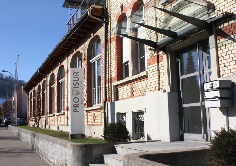 Außenansicht des Firmengebäudes der Provisur Technologies GmbH in Flawil aus einer seitlichen Perspektive bei Sonnenschein. Zu sehen ist eine traditionelle Backsteinfassade mit großen Bogenfenstern, einem modernen Glaseingang mit Briefkästen und einer weißen Stehle mit dem Firmenlogo „Provisur Technologies“.