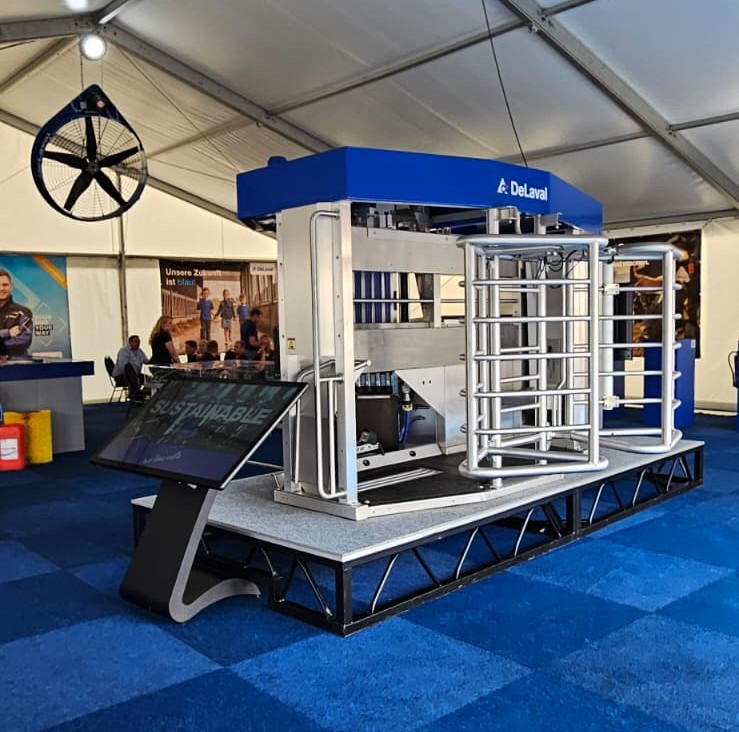 Totale eines DeLaval-Ausstellungsstandes in einem großen Veranstaltungszelt. Auf einem erhöhten Podest ist ein automatisches Melksystem (VMS) mit blauem Dach und silbernem Schutzgitter aufgebaut. Links davor steht eine schwarze diliton-Infostele mit einem schrägen Touch-Bildschirm, auf dem das Wort „SUSTAINABLE“ zu sehen ist. Der Hallenboden ist mit einem blau karierten Teppich ausgelegt.
