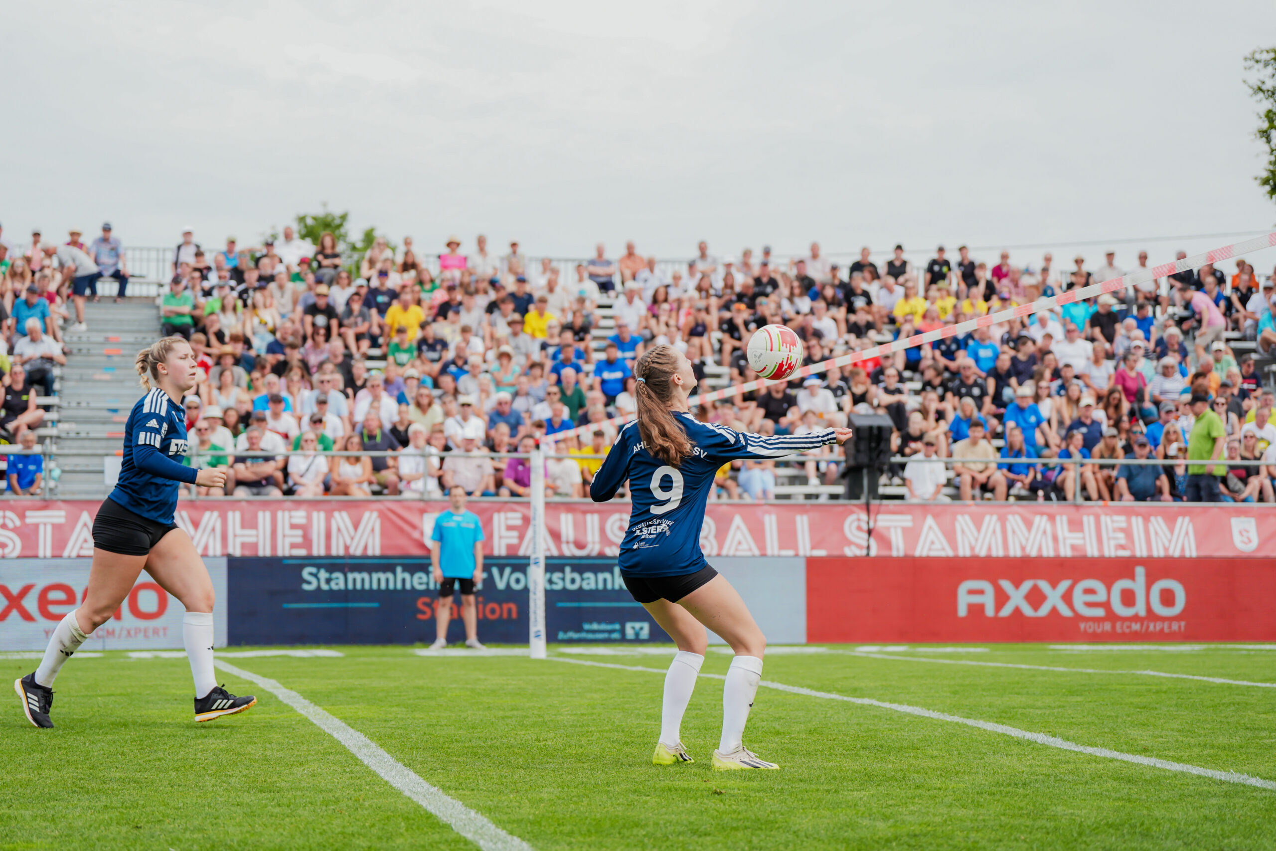 Eine Faustballspielerin mit der Rückennummer 9 nimmt den Ball während des Frauen-Viertelfinals der Deutschen Meisterschaft 2024 an. Fotografiert von Lars Neumann am 27. Juli 2024 beim Spiel des Ahlhorner SV gegen den TSV Pfungstadt im Stammheimer Volksbank-Stadion. Links eilt eine Teamkollegin herbei, während im Hintergrund gut gefüllte Zuschauerränge auf einer Tribüne und rote Bandenwerbung zu sehen sind.