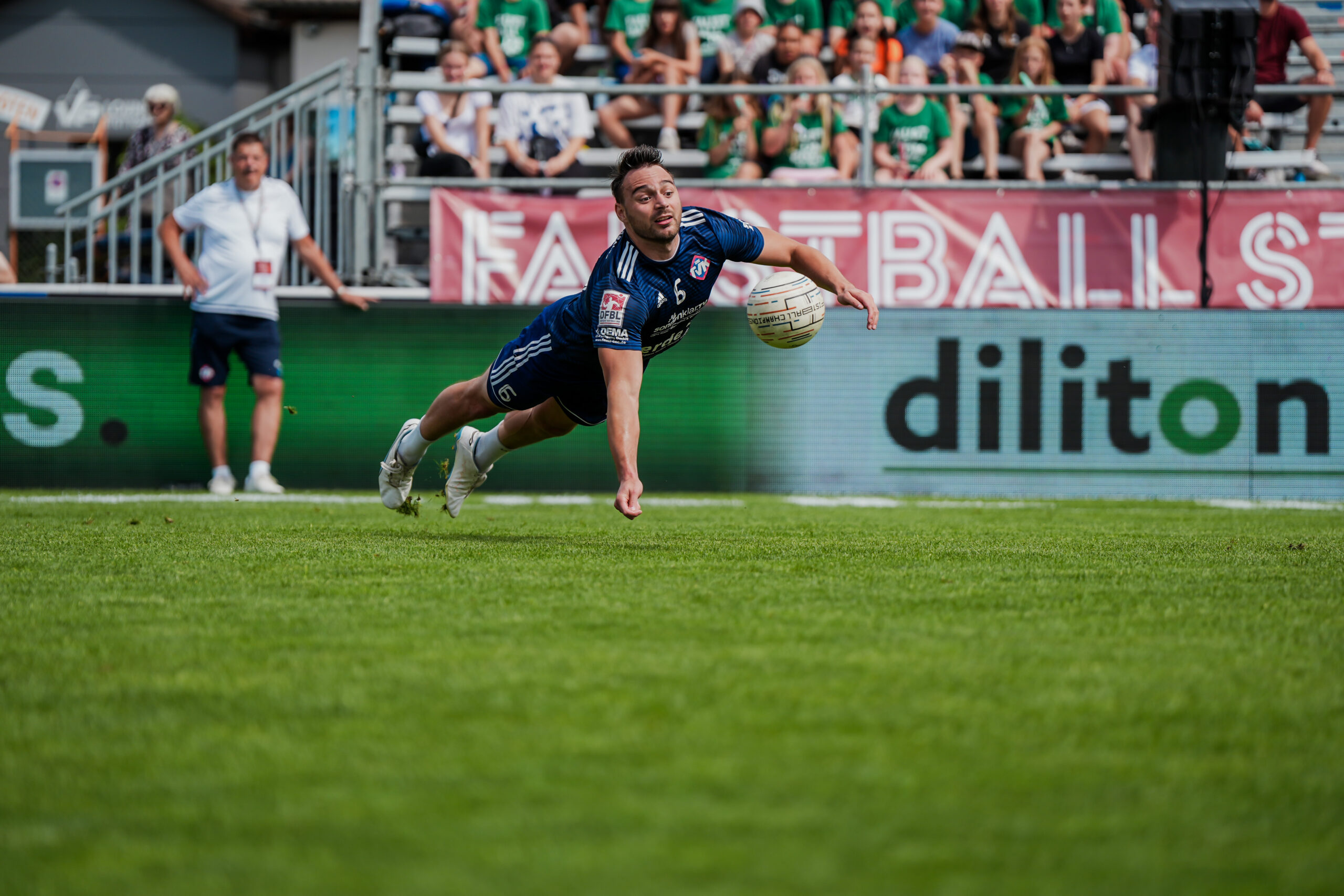 Ein Faustballspieler im blauen Trikot mit der Nummer 6 hechtet im vollen Körpereinsatz parallel zum Rasen, um den Ball mit der Faust abzuwehren. Action-Aufnahme von Fotograf Lars Neumann beim Männer-Viertelfinale der Deutschen Meisterschaft 2024 zwischen dem TV Brettorf und dem TV Käfertal am 27. Juli 2024. Im Hintergrund sind Zuschauer auf einer Tribüne und Bandenwerbung zu sehen.