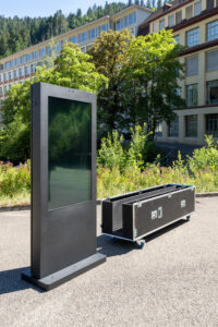 Das Bild zeigt eine freistehende, moderne digitale Outdoor-Stele in edlem Mattschwarz auf einem asphaltierten Platz. Der vertikal ausgerichtete, wetterfeste Bildschirm ist ausgeschaltet und spiegelt die grüne Umgebung leicht wider. Rechts hinter der Stele steht das maßgeschneiderte, schwarze Transport-Case, auch Flightcase genannt, mit silbernen Metallecken, Butterfly-Verschlüssen und blauen Lenkrollen. Das Case ist nach oben hin geöffnet und bereit für das sichere Verstauen des Bildschirms. Im Hintergrund ist ein helles Gebäude mit großen Fenstern und grünen Bäumen zu sehen.