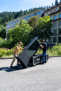 Das Bild zeigt zwei Männer im Außenbereich, die gemeinsam den großen, schwarzen digitalen Outdoor-Kundenstopper anheben, um ihn schräg in das am Boden stehende, schwarze Flightcase hineinzuführen oder herauszuheben. Der Mann links trägt eine gelbe Jacke und helle Hosen, der Mann rechts ein schwarzes T-Shirt, Jeans und eine Sonnenbrille. Die Szene verdeutlicht die Handhabung beim Transport des Event-Equipments auf einem asphaltierten Platz vor einem großen Fabrikgebäude und bewaldeten Bergen im Hintergrund.