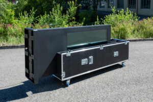Das Bild zeigt den schwarzen digitalen Outdoor-Kundenstopper in einer horizontalen Kippposition im Außenbereich. Die Stele ist halbseitig in die untere Schale des geöffneten Flightcases eingelegt, während die linke Seite (der Standfuß der Stele) noch leicht herausragt und die Montagepunkte an der Unterseite sichtbar macht. Das schwarze Case steht auf blauen Lenkrollen und ist mit silbernen Metallecken und Verschlüssen versehen. Im Hintergrund befinden sich grüne Büsche und ein helles Gebäude.