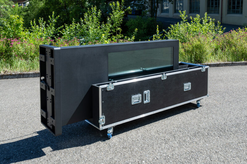 Das Bild zeigt den schwarzen digitalen Outdoor-Kundenstopper in einer horizontalen Kippposition im Außenbereich. Die Stele ist halbseitig in die untere Schale des geöffneten Flightcases eingelegt, während die linke Seite (der Standfuß der Stele) noch leicht herausragt und die Montagepunkte an der Unterseite sichtbar macht. Das schwarze Case steht auf blauen Lenkrollen und ist mit silbernen Metallecken und Verschlüssen versehen. Im Hintergrund befinden sich grüne Büsche und ein helles Gebäude.