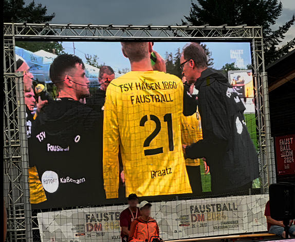 Große LED-Videowall bei der Deutschen Faustball-Meisterschaft 2024 im Stadion. Auf dem Bildschirm ist eine Spielerszene des TSV Hagen 1860 mit der Trikotnummer 21 (Braatz) und Betreuern zu sehen. Unter der Leinwand hängt ein Banner mit der Aufschrift Faustball DM 2024.