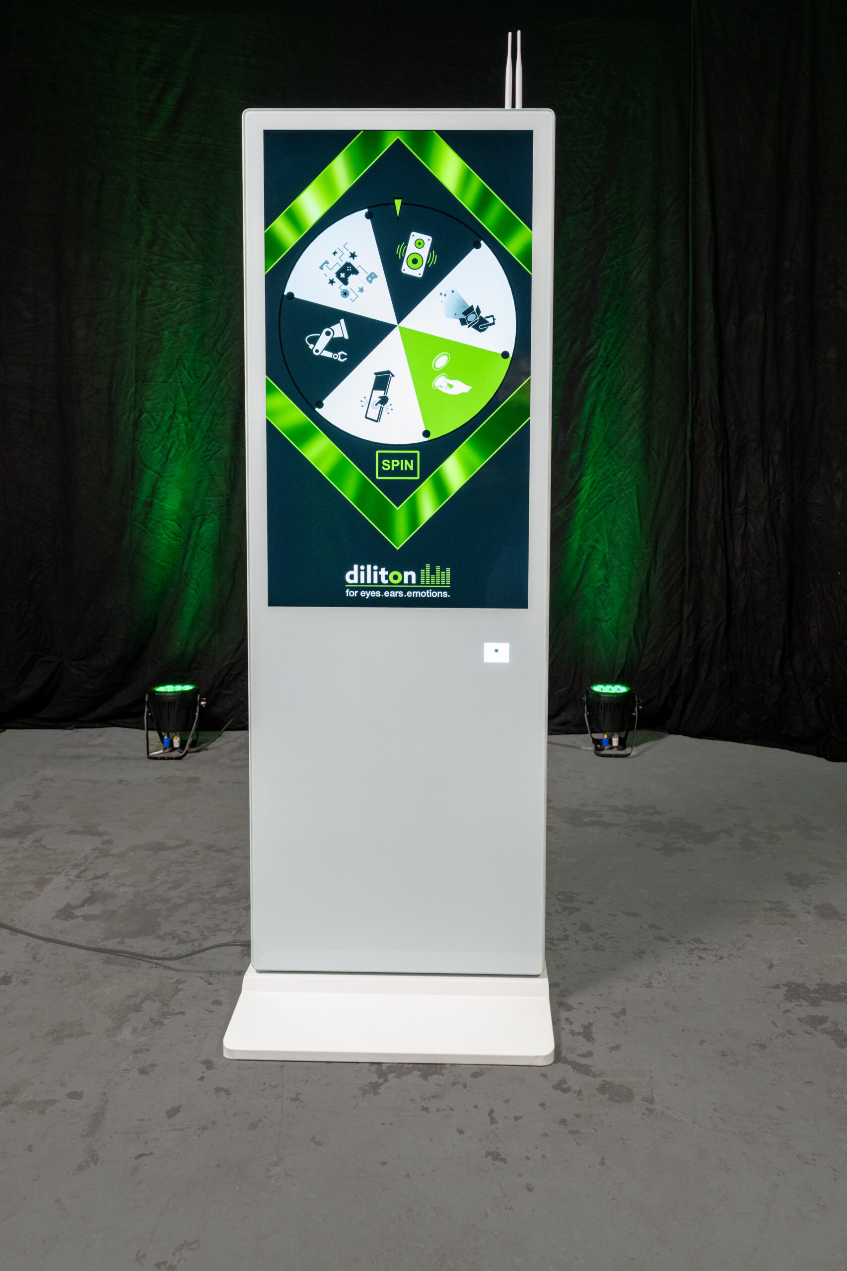 Frontansicht einer freistehenden, weißen Touch-Stele im Hochformat auf grauem Boden. Das große vertikale Display zeigt ein interaktives, grün-weißes Glücksrad mit verschiedenen Symbolen und einem „SPIN“-Button im Zentrum sowie das Logo „diliton“ am unteren Bildschirmrand. Rechts unter dem Display ist das quadratische Fenster des integrierten und optional zubuchbaren QR-Code-Scanners platziert und am oberen Gehäuserand sind zwei kleine Antennen sichtbar. Diese Digital Signage Stele dient als digitaler Kundenstopper vor einem dunklen Hintergrund mit grüner Scheinwerferbeleuchtung.