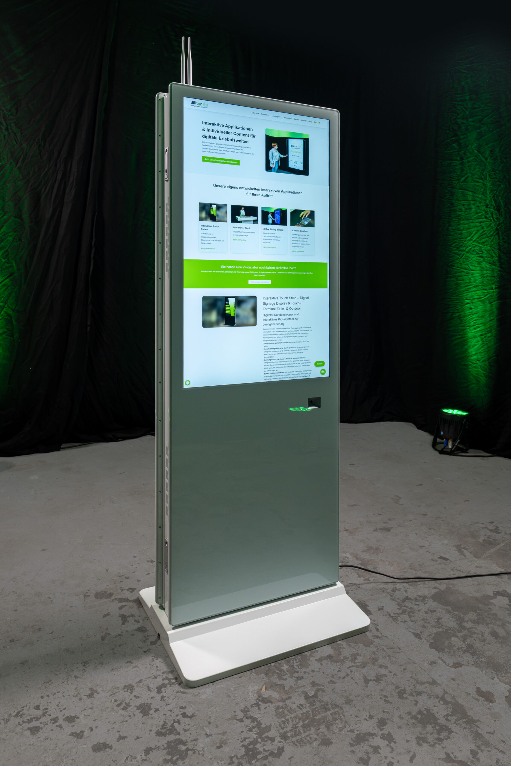 Perspektivische Frontansicht einer freistehenden, weißen Touch-Stele im Hochformat auf grauem Betonboden. Das große vertikale Display zeigt eine hell gestaltete Webseite mit Textblöcken und Bildern in Grüntönen. Rechts unter dem Bildschirm ist das quadratische Fenster des integrierten und optional zubuchbaren QR-Code-Scanners zu sehen, am oberen Rand ragen zwei WLAN-Antennen auf. Das schlanke Gehäuse dieser Digital Signage Stele steht leicht schräg auf einem weißen Sockel vor einem dunklen Hintergrund mit grüner Effektbeleuchtung.