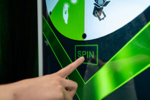 Nahaufnahme einer Nutzerinteraktion auf dem Display einer weißen Digital Signage Stele. Ein Finger berührt ein grünes, quadratisches Feld mit der Aufschrift „SPIN“ auf dem Touchscreen. Darüber ist ein Ausschnitt eines digital dargestellten Glücksrads in den Farben Grün und Weiß zu sehen. Diese interaktive Anwendung verdeutlicht die Nutzung der Touch-Stele als digitaler Kundenstopper für Gewinnspiele oder Messen.