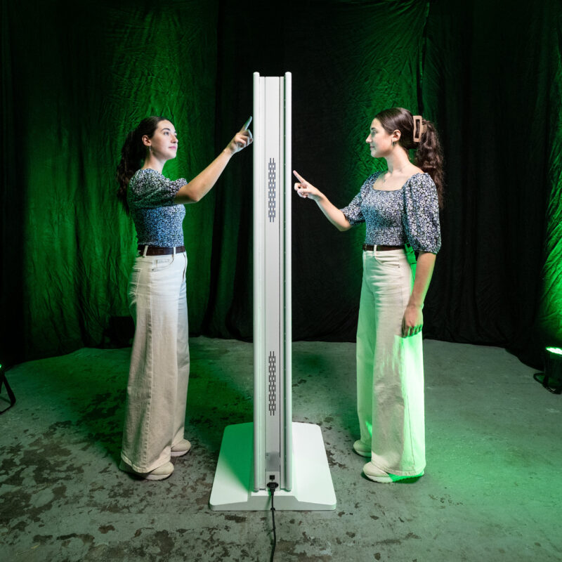 Totale Seitenansicht einer weißen, doppelseitigen Touch-Stele zur Veranschaulichung der beidseitigen Nutzung. Links und rechts der schlanken Digital Signage Stele steht jeweils eine junge Frau mit dunklen Haaren, gemusterter Bluse und heller Hose im Profil. Beide interagieren gleichzeitig per Fingertipp mit den gegenüberliegenden Bildschirmen. Das Gehäuse zeigt Lüftungsschlitze an der Seite und steht auf einem weißen Sockel. Der dunkle Hintergrund wird von Scheinwerfern grün ausgeleuchtet.