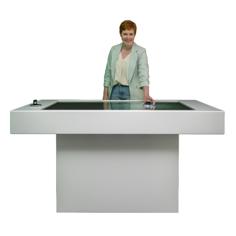 Das Bild zeigt eine Frau mit kurzen, rötlichen Haaren und einer hellen Jacke, die lächelnd hinter einem großen, modernen Multi-Touch-Tisch steht. Der Tisch besitzt ein minimalistisches, weißes Gehäuse und einen breiten Standfuß. Die Frau platziert ihre linke Hand auf einem physischen Objektmarker auf der Display-Oberfläche, um digitale Inhalte interaktiv zu steuern. Am linken Rand des Tisches ist ein weiteres Bedienobjekt aufgesetzt.