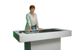 Das Bild zeigt aus einer seitlichen Perspektive eine Frau mit kurzen, rötlichen Haaren und einer hellen Jacke an dem modernen, weißen Multi-Touch-Tisch. Sie blickt leicht nach unten konzentriert auf die Bildschirmoberfläche. Ihre rechte Hand bewegt einen runden Objektmarker im linken Bereich des Displays, während ihre linke Hand flach auf dem Rahmen des Tisches ruht. Am äußeren linken Rand ist ein weiteres, Steuerungsobjekt aufgesetzt.