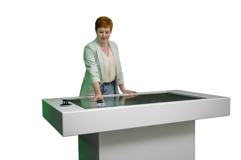 Das Bild zeigt aus einer seitlichen Perspektive eine Frau mit kurzen, rötlichen Haaren und einer hellen Jacke an dem modernen, weißen Multi-Touch-Tisch. Sie blickt leicht nach unten konzentriert auf die Bildschirmoberfläche. Ihre rechte Hand bewegt einen runden Objektmarker im linken Bereich des Displays, während ihre linke Hand flach auf dem Rahmen des Tisches ruht. Am äußeren linken Rand ist ein weiteres, Steuerungsobjekt aufgesetzt.