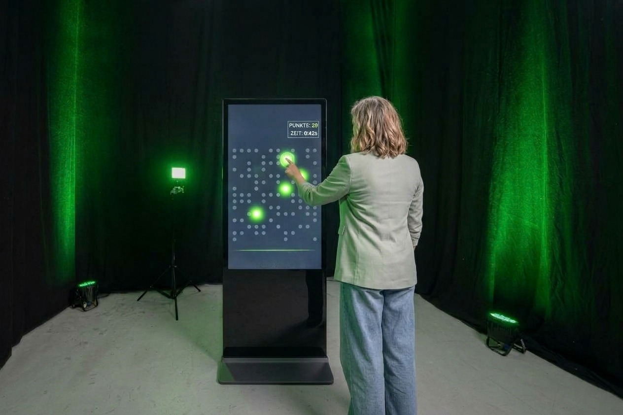 Eine Frau bedient ein interaktives Reaktionsspiel auf einer digitalen Touch-Stele, bei dem sie leuchtende Punkte auf dem Bildschirm berührt.